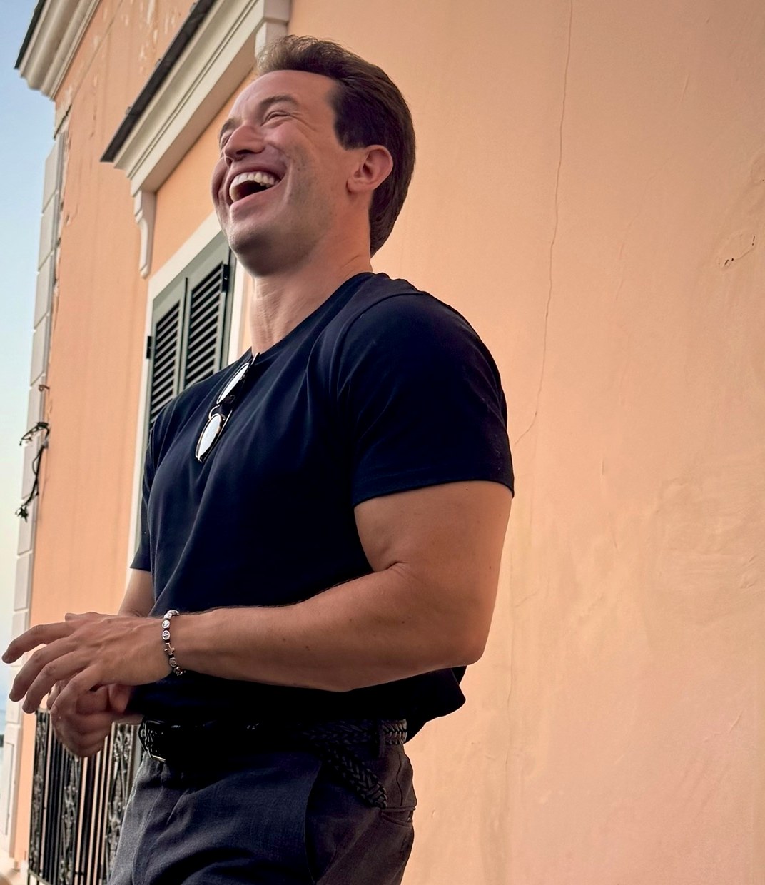 Michael Hewitt laughing on a terrace at sunset, in a black T-shirt against a peach wall