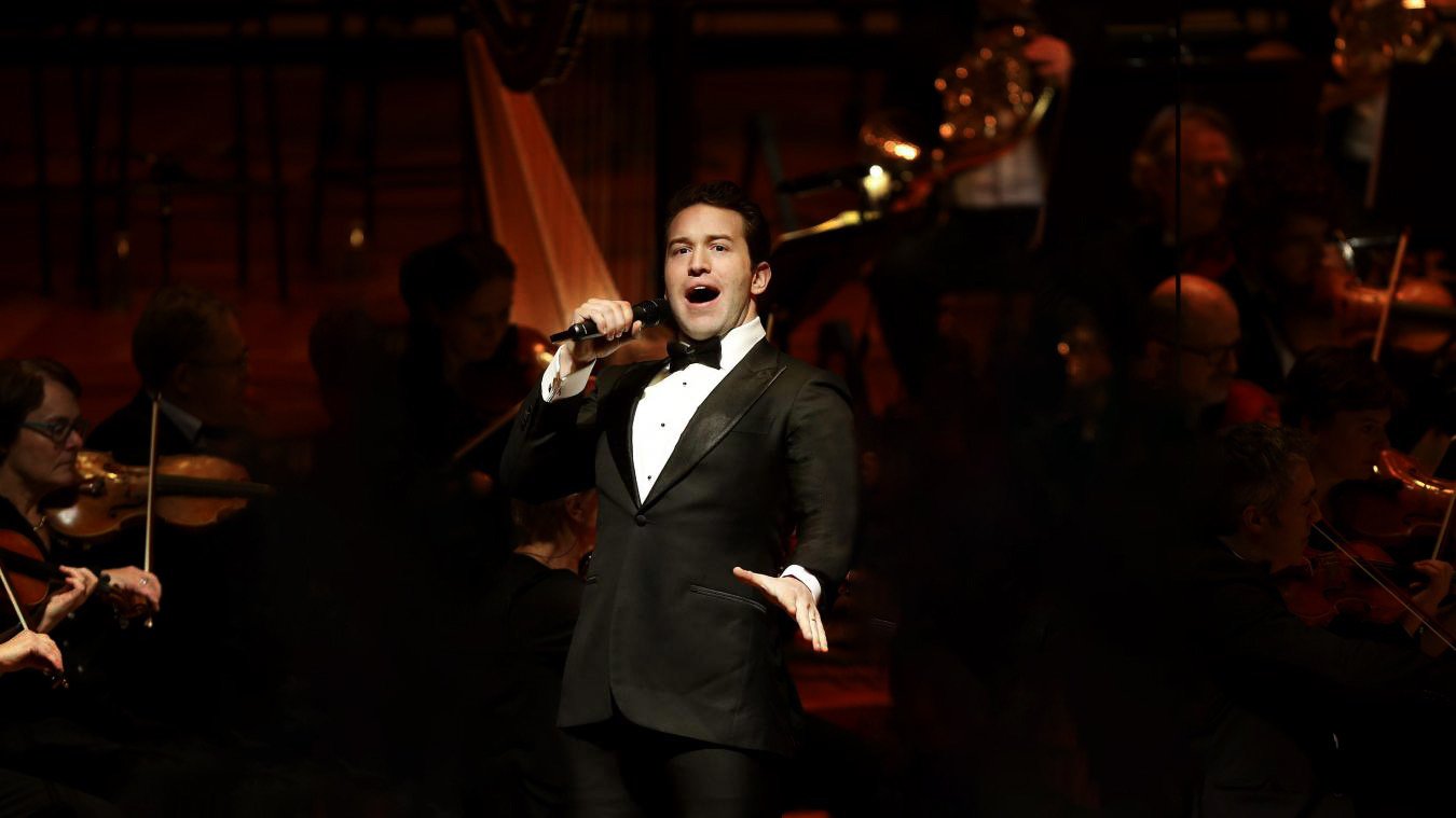 Michael Hewitt in a tuxedo singing into a microphone in front of a full orchestra