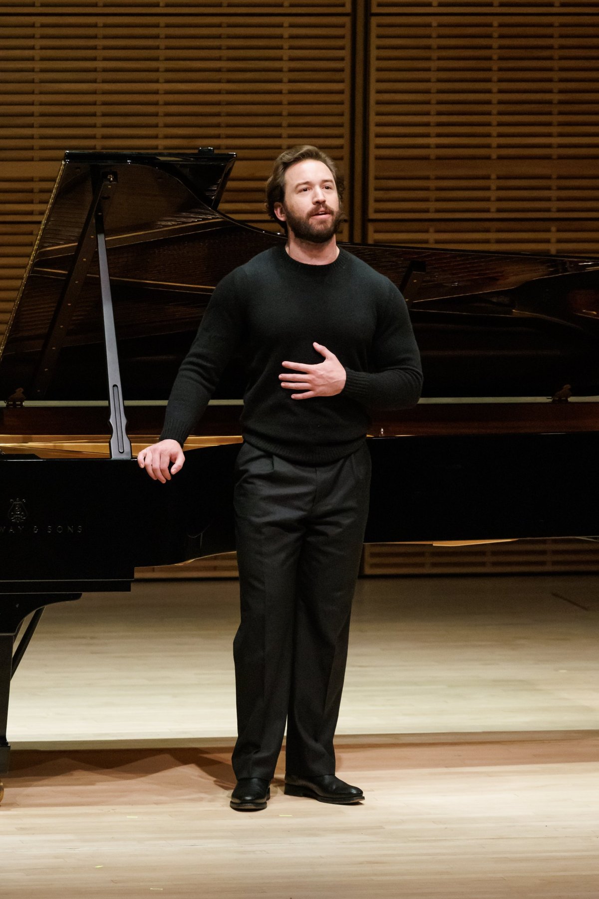 Michael Hewitt in a black sweater, singing beside a grand piano in a wood-panelled recital hall